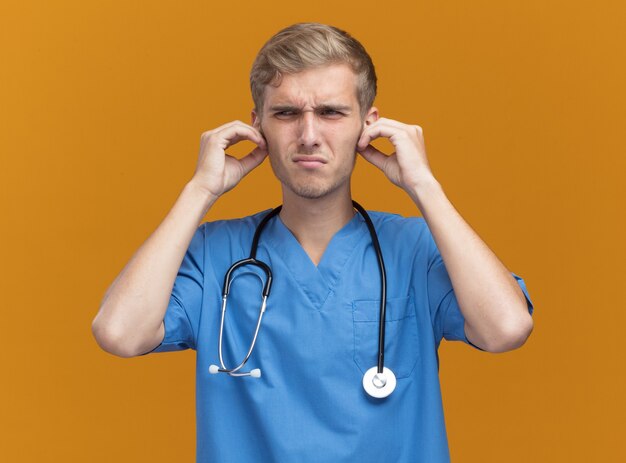 Insatisfait jeune médecin de sexe masculin portant l'uniforme de médecin avec stéthoscope a attrapé les oreilles isolé sur mur orange