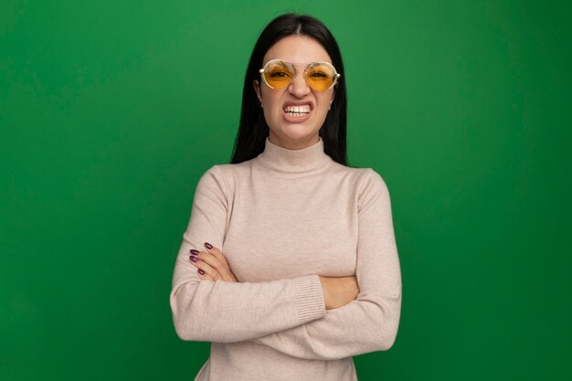 Insatisfait jolie fille caucasienne brune à lunettes de soleil se dresse avec les bras croisés sur le vert