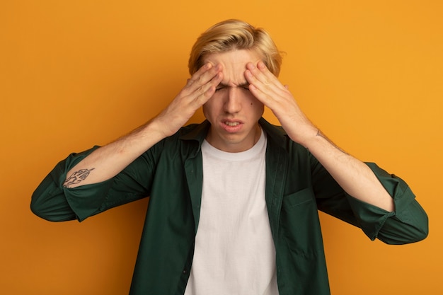 Insatisfait avec les yeux fermés jeune mec blond portant un t-shirt vert mettant les mains sur le front