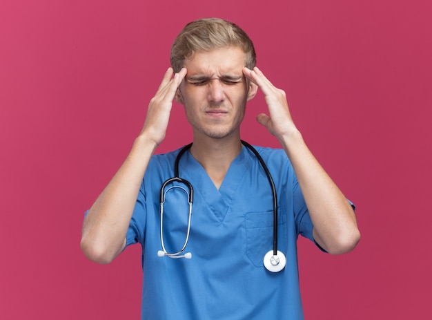 Insatisfait les yeux fermés jeune médecin de sexe masculin portant l'uniforme de médecin avec stéthoscope mettant les doigts sur les tempes douloureuses isolé sur mur rose