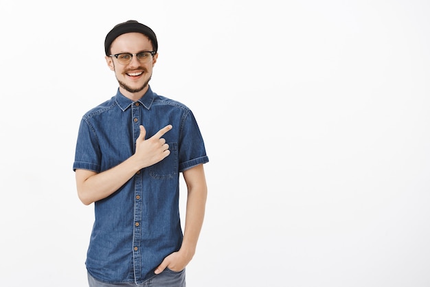 Insouciant heureux et satisfait beau photographe masculin en bonnet noir à la mode et chemise bleue pointant vers le coin supérieur droit souriant largement de satisfaction et de plaisir