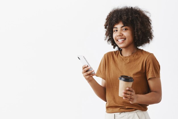 Insouciante détendue et joyeuse jeune fille à la peau sombre avec une coiffure afro en t-shirt marron debout à moitié tourné avec un smartphone et une tasse en papier de café dans les mains, la messagerie ou la navigation sur Internet