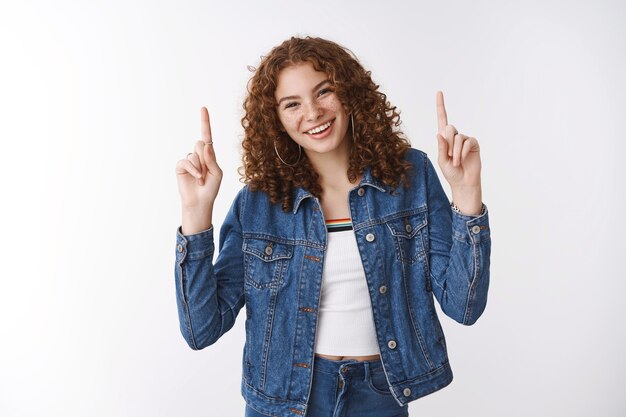 Insouciante heureuse charmante souriante rousse européenne taches de rousseur peau post-acné riant joyeusement lever les mains pointant l'index vers le haut promouvoir la publicité du produit debout ravi mur blanc optimiste