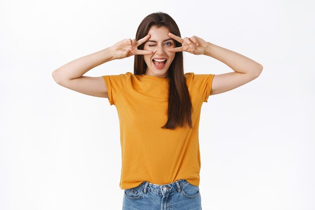 Insouciante impertinente heureuse jeune femme brune en t-shirt jaune montrant la paix ou le signe de la victoire sur les yeux clignotant joyeusement souriant montrant la langue profiter d'une fête géniale s'amusant fond blanc