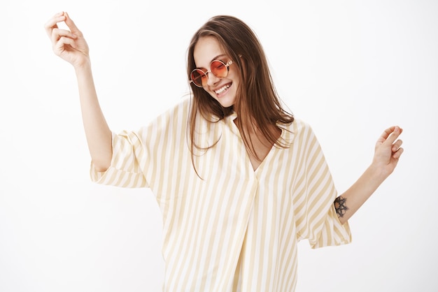 Insouciante ravie et heureuse jeune femme moderne dans des lunettes de soleil rouges élégantes et chemisier rayé jaune danse féminine et joyeuse souriant de joie s'amuser