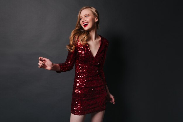 Inspiré de la jeune femme en robe rouge dansant à la fête. Photo de Studio de fille blonde heureuse posant sur fond sombre.