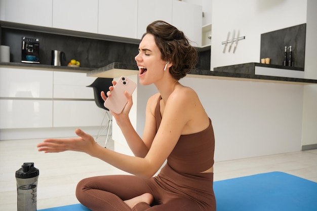 Photo gratuite un instructeur d'entraînement de fitness insouciant est assis à la maison avec une bouteille d'eau, un tapis de yoga porte un survêtement sin