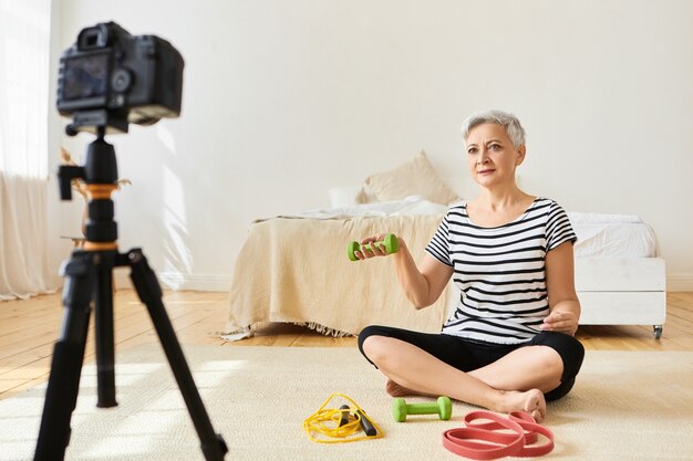 Instructeur de fitness féminin senior athlétique avec des cheveux gris courts exerçant sur le sol avec des haltères verts, enregistrant un didacticiel vidéo via une caméra sur un trépied. Personnes, âge et mode de vie sain et actif