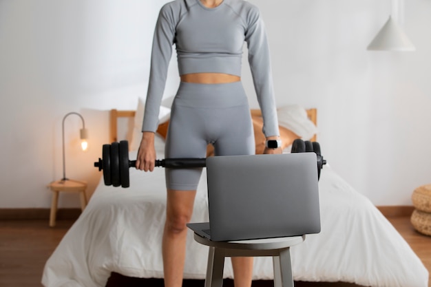 Instructeur de fitness féminin soulevant des poids à la maison devant un ordinateur portable