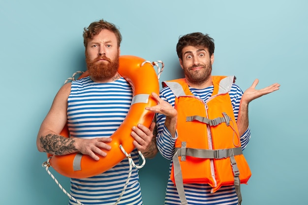 Instructeur de natation masculin sérieux avec bouée de sauvetage, stagiaire douteux porte un gilet orange, écarte les mains