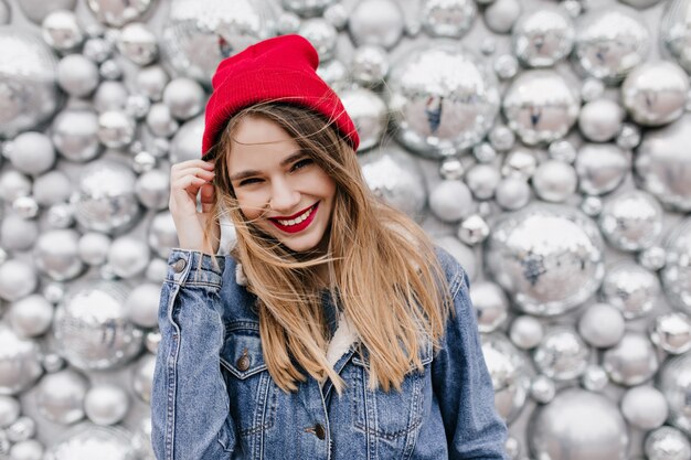 Intéressé belle fille en veste en jean touchant ses longs cheveux. Sourire modèle féminin européen en chapeau posant sur un mur brillant.