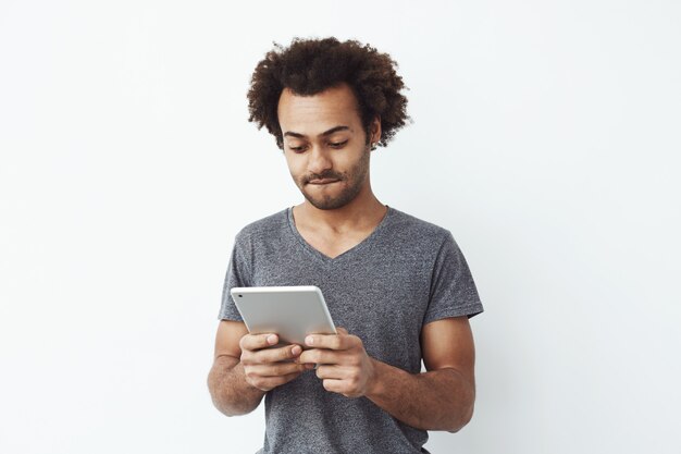 Intéressé et concentré jeune homme africain regardant tablette jouer à un jeu de plateforme et profiter de nouveaux niveaux sur fond blanc.