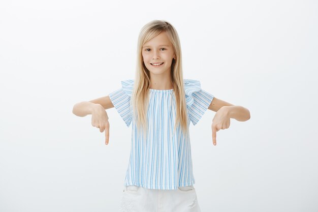 Intéressé heureux jeune fille aux cheveux blonds, pointant vers le bas avec l'index et souriant largement, étant sûr de lui et détendu sur un mur gris