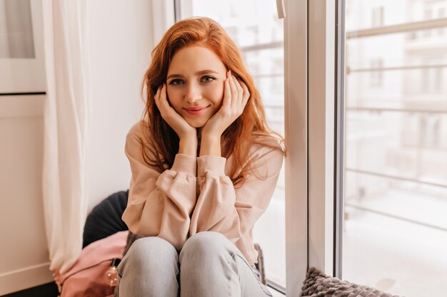 Intéressée jolie fille en jeans touchant son visage. Ginger joyeuse dame posant dans son appartement.