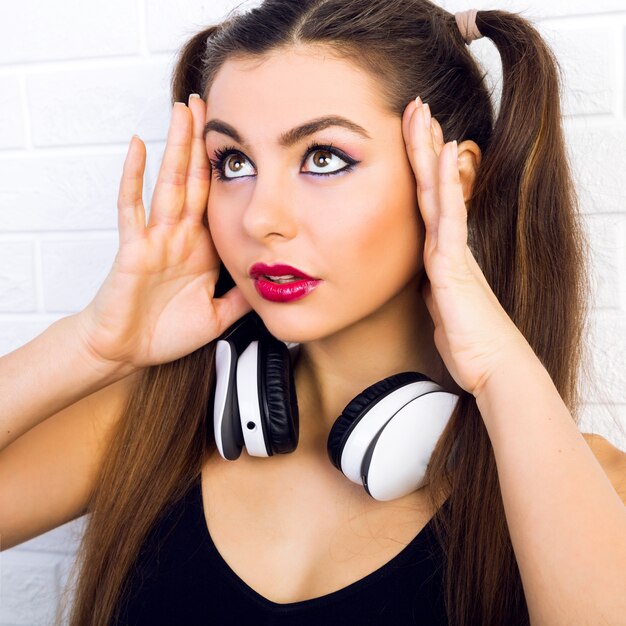 Intérieur gros plan portrait de mode de belle jeune femme, avec maquillage et coiffure à la mode, écoute de la musique dans les écouteurs, portrait lumineux urbain de fille sexy dj