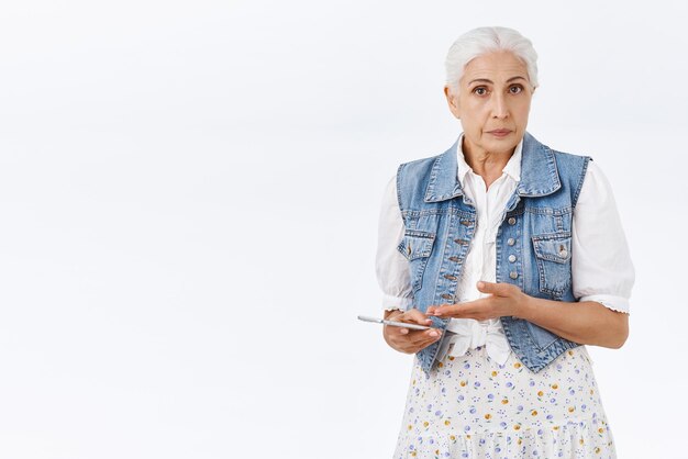 Interrogée et bouleversée, une vieille femme âgée ne peut pas comprendre comment se connecter au courrier à l'aide d'un smartphone, tenir un téléphone portable pointant confus vers l'appareil et regarder la caméra perplexe et indécise