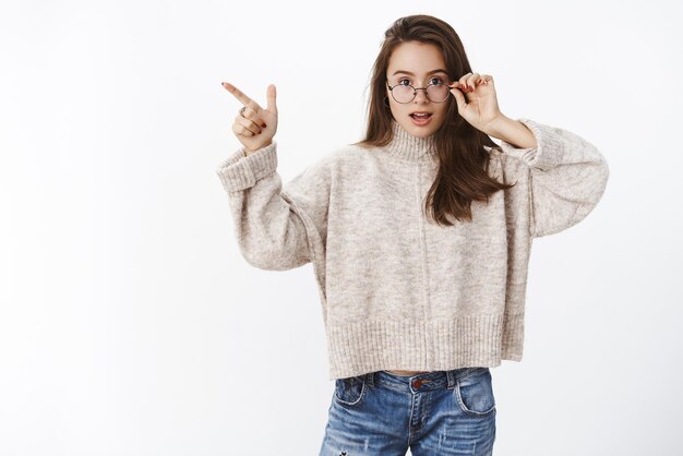 Interrogée et impressionnée jolie jeune femme de 20 ans qui enlève des lunettes intriguée en pointant vers le coin supérieur gauche étant curieuse avec le produit posant une question à la caméra sur fond gris