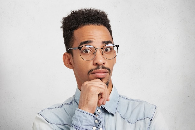 Intrigué et confus, bel homme à la peau sombre avec une coiffure afro garde la main sur le menton, regarde dans la confusion