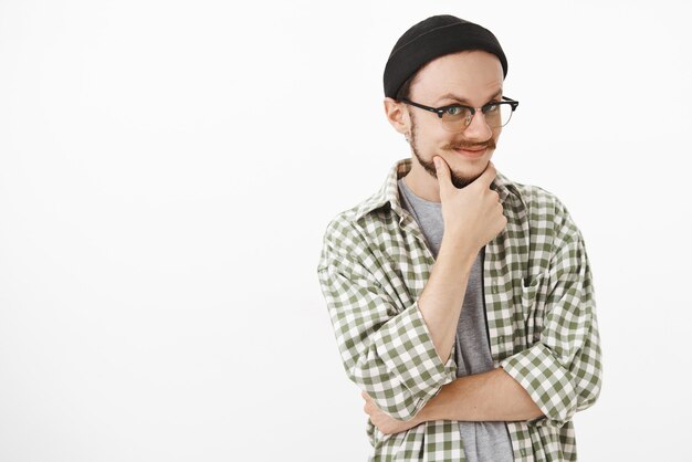 intrigué enthousiaste jeune homme créatif à lunettes et bonnet noir frottant la barbe souriant de curiosité et de joie regardant mystérieusement comme s'il avait une idée ou une intention