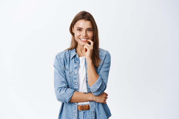 Intrigué jeune femme touchant la lèvre et souriant en voyant quelque chose d'intéressant à la recherche réfléchie et heureuse debout sur fond blanc