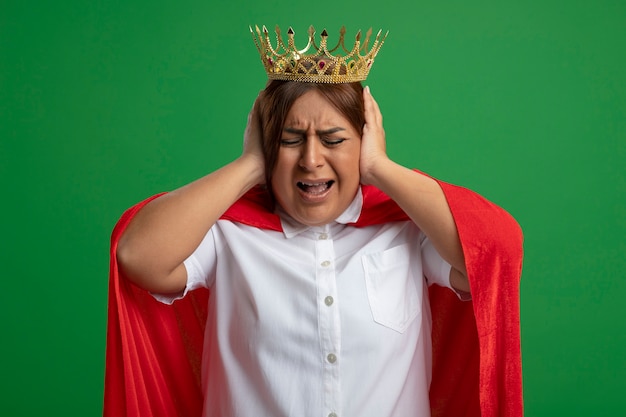 Irritant femme de super-héros d'âge moyen portant couronne mettant les mains sur les oreilles isolé sur fond vert