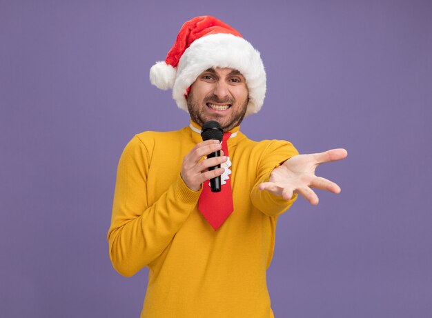 Irrité jeune homme de race blanche portant chapeau de Noël et cravate tenant le microphone regardant la caméra étendant la main vers la caméra isolée sur fond violet