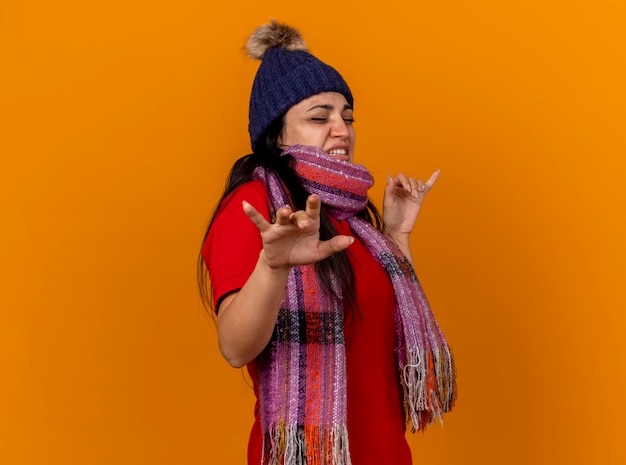 Photo gratuite irritée jeune femme malade portant un chapeau d'hiver et une écharpe debout en vue de profil en gardant les mains en l'air avec les yeux fermés isolé sur un mur orange