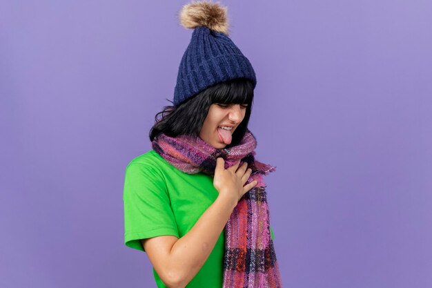 Irritée jeune femme malade portant un chapeau d'hiver et une écharpe en gardant la main sur la poitrine montrant la langue avec les yeux fermés isolé sur mur violet avec copie espace