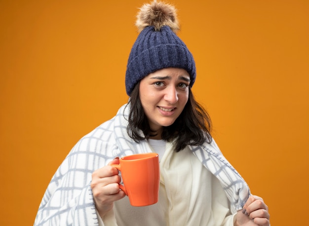 Irritée jeune femme malade portant robe chapeau d'hiver enveloppé dans un plaid tenant une tasse de thé à l'avant saisissant plaid isolé sur mur orange