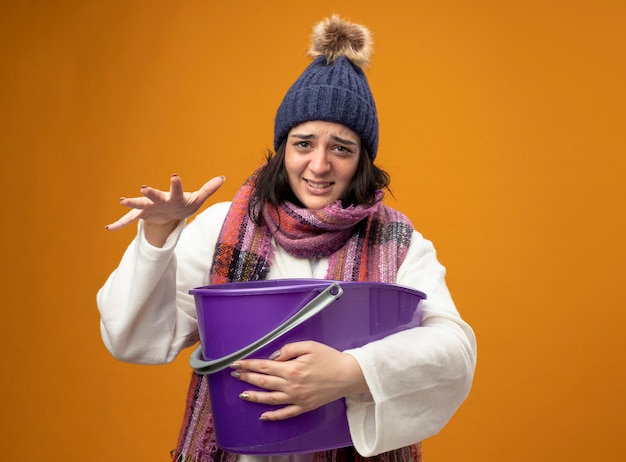 Irritée jeune fille malade de race blanche portant chapeau d'hiver robe et écharpe ayant des nausées tenant un seau en plastique gardant la main dans l'air isolé sur un mur orange avec copie espace