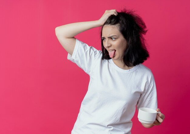 Irritée jeune jolie fille caucasienne tenant la tasse à côté et mettant la main sur la tête isolée sur fond cramoisi avec espace de copie