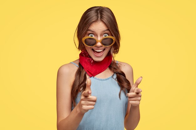 J'ai de beaux projets sur toi. Une fille joyeuse à la mode pointe avec les deux index directement sur quelque chose de merveilleux, porte des lunettes de soleil à la mode, isolées sur un mur jaune.