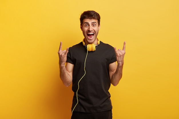 J'aime le heavy metal! Joyeux jeune homme étant fou, fait un geste de corne avec les deux mains, écoute la musique préférée dans les écouteurs