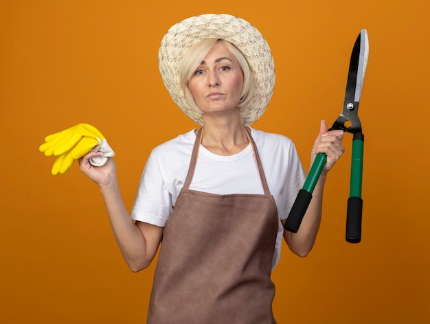 Photo gratuite jardinier d'âge moyen confiant femme en uniforme de jardinier portant un chapeau tenant des gants de jardinier et des cisailles à haie