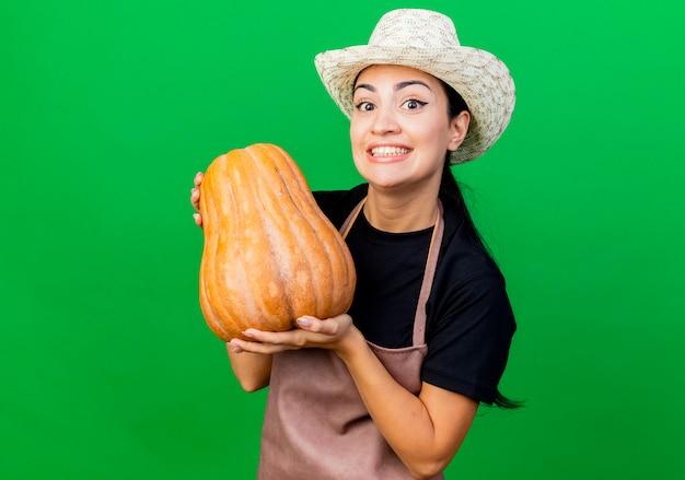 Photo gratuite jardinier de belle jeune femme en tablier et chapeau tenant la citrouille souriant