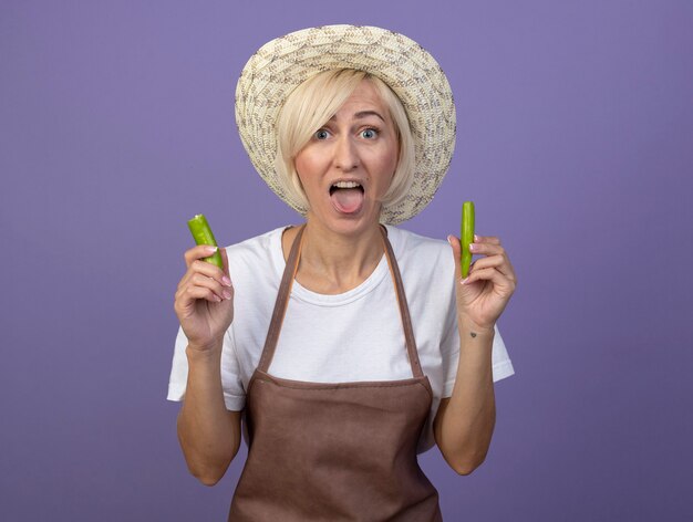 Jardinier blonde d'âge moyen ludique femme en uniforme portant un chapeau tenant des moitiés de poivre montrant la langue
