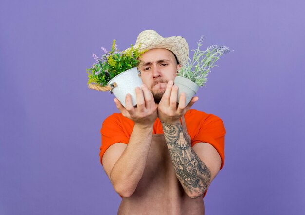 Photo gratuite jardinier mâle confus portant chapeau de jardinage détient des fleurs dans des pots de fleurs