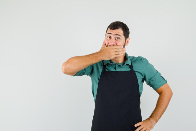 Jardinier masculin couvrant la bouche avec la main en t-shirt, tablier et l'air surpris, vue de face.