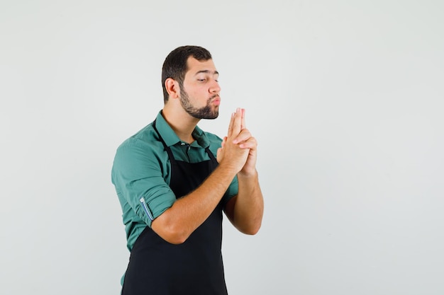 Jardinier masculin en t-shirt, tablier soufflant sur un pistolet fabriqué à la main et ayant l'air confiant, vue de face.