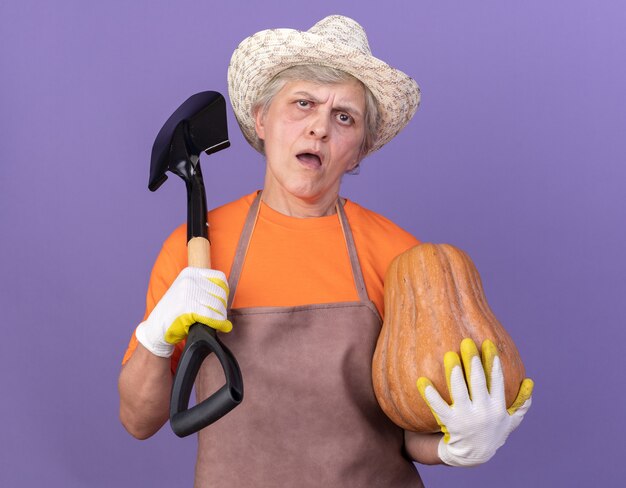 Jardinière âgée agacée portant un chapeau et des gants de jardinage tenant une citrouille et une pelle sur l'épaule