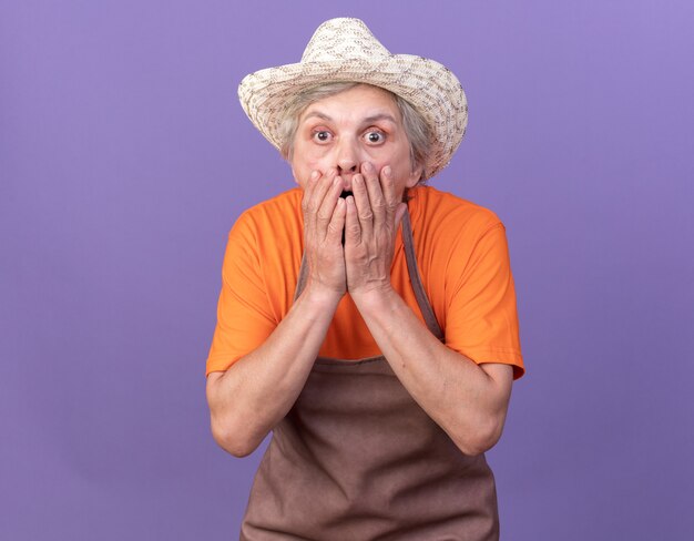 Une jardinière âgée choquée portant un chapeau de jardinage met les mains sur la bouche isolée sur un mur violet avec espace pour copie