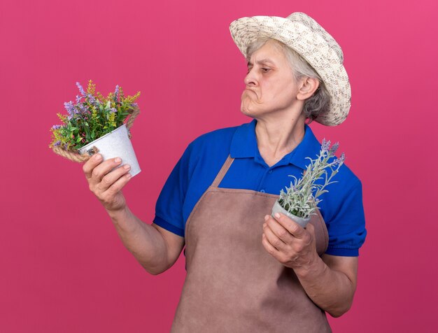 Une jardinière âgée impressionnée portant un chapeau de jardinage tenant et regardant des pots de fleurs