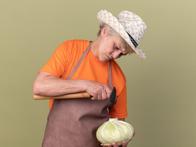 Jardinière âgée sans idée portant un chapeau de jardinage tenant un râteau et regardant le chou isolé sur un mur vert olive avec espace pour copie