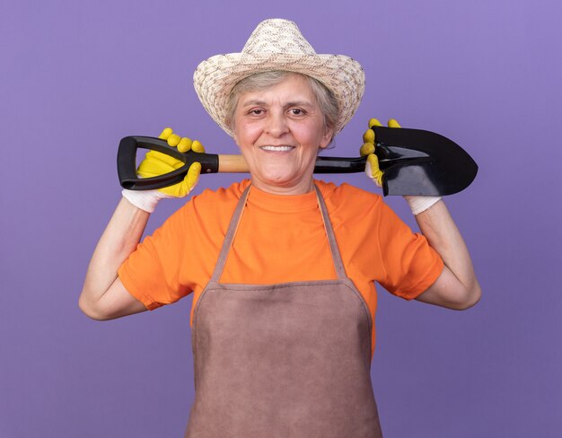 Jardinière âgée souriante portant un chapeau de jardinage et des gants tenant une pelle sur le cou derrière isolé sur un mur violet avec espace pour copie