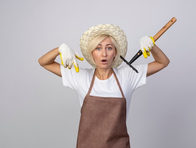 Une jardinière blonde d'âge moyen impressionnée en uniforme portant un chapeau et des gants de jardinage tenant un râteau pointant vers le bas