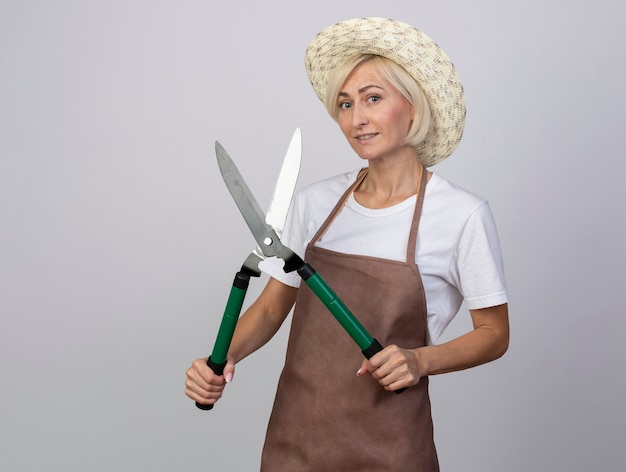 Jardinière blonde d'âge moyen souriante en uniforme portant un chapeau tenant des cisailles à haies isolées sur un mur blanc avec espace pour copie