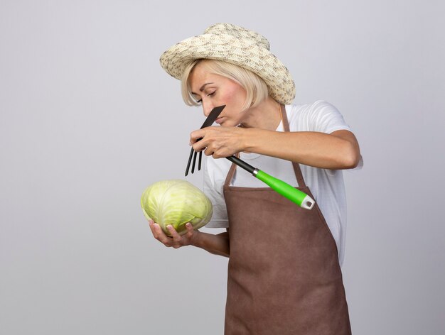Jardinière blonde d'âge moyen en uniforme portant un chapeau tenant du chou et un râteau au-dessus en regardant le chou isolé sur un mur blanc avec espace pour copie