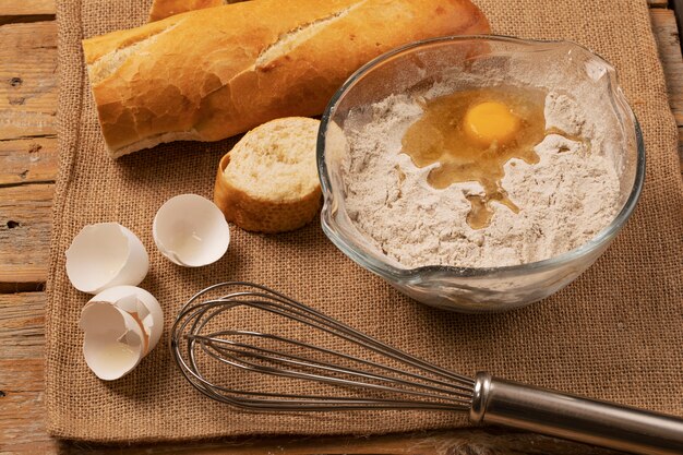 Jaune d'oeuf sur la farine. Coquilles d'oeuf, tranches de baguette et batteur à main sur un morceau de toile de jute.