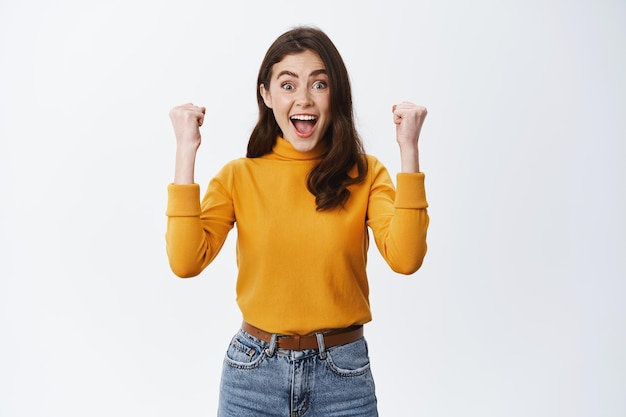 Je l'ai fait. Fille excitée se réjouissant, levant les mains et souriant heureux, atteindre l'objectif ou le prix, gagner de l'argent et célébrer, debout dans des vêtements décontractés sur fond blanc