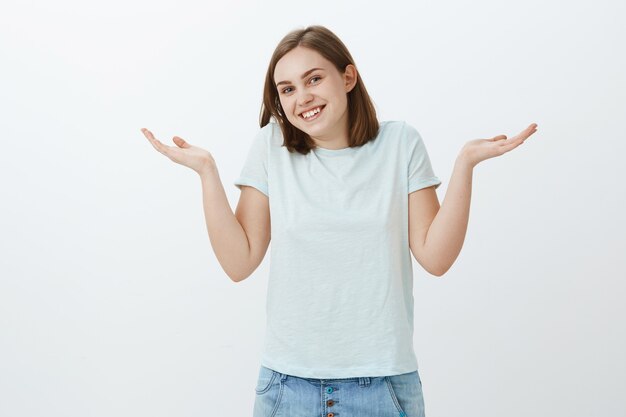 Je n'ai aucune idée et je m'en fiche. Chill et heureuse femme insouciante haussant les épaules avec les mains écartées près des épaules souriant joyeusement étant ignorant et inconscient debout indifférent sur mur gris